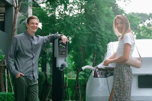 Young couple travel with EV electric car charging in green sustainable city outdoor garden in summer shows urban sustainability lifestyle by green clean rechargeable energy of electric vehicle innards