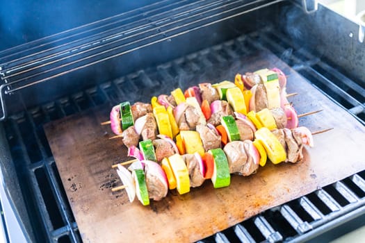 Skewered beef and fresh veggies sizzle on a copper grill mat over a gas outdoor grill.