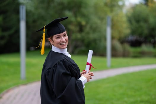 Portrait of happy caucasian woman in graduate gown holding diploma outdoors