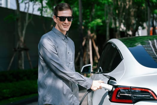 Young man travel with EV electric car charging in green sustainable city outdoor garden in summer shows urban sustainability lifestyle by green clean rechargeable energy of electric vehicle innards