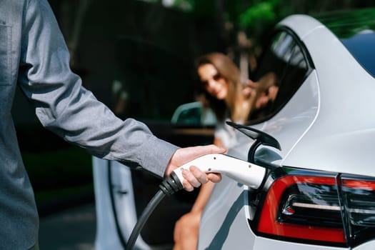 Young couple travel with EV electric car charging in green sustainable city outdoor garden in summer shows urban sustainability lifestyle by green clean rechargeable energy of electric vehicle innards