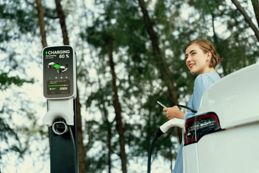Holiday road trip vacation by the green countryside nature with beautiful young woman recharging electric vehicle with alternative energy. Environmental friendly travel wit EV car. Perpetual