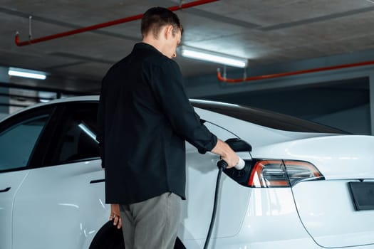 Young man travel with EV electric car to shopping center parking lot charging in downtown city showing urban sustainability lifestyle by green clean rechargeable energy of electric vehicle innards