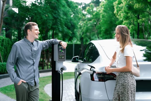 Young couple travel with EV electric car charging in green sustainable city outdoor garden in summer shows urban sustainability lifestyle by green clean rechargeable energy of electric vehicle innards