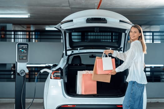 Young woman travel with EV electric car to shopping center parking lot charging in downtown city showing urban sustainability lifestyle by green clean rechargeable energy of electric vehicle innards