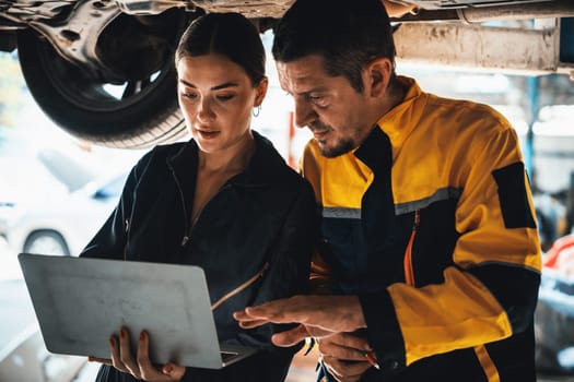 Two vehicle mechanic working together underneath lifted car, conduct car inspection with laptop. Automotive service technician in uniform carefully make diagnostic troubleshooting. Oxus