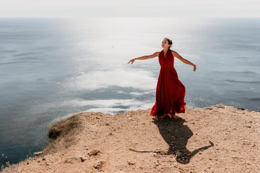 Side view a Young beautiful sensual woman in a red long dress posing on a rock high above the sea during sunrise. Girl on the nature on blue sky background. Fashion photo.