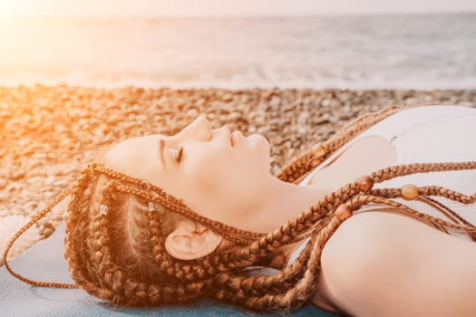 Woman yoga sea. Well looking middle aged woman with braids dreadlocks in white leggings and tops doing stretching pilates on yoga mat near sea. Female fitness yoga routine concept. Healthy lifestyle