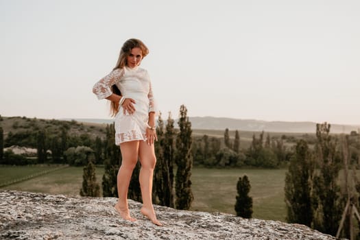 Romantic beautiful bride in white dress posing with sea and mountains in background. Stylish bride standing back on beautiful landscape of sea and mountains on sunset