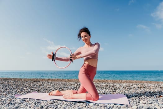 Middle aged well looking woman with black hair doing Pilates with the ring on the yoga mat near the sea on the pebble beach. Female fitness yoga concept. Healthy lifestyle, harmony and meditation.