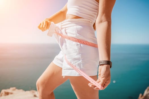 a young and slender woman in shorts holds in her hand a measuring yellow tape on her sexy thighs on the beach. concept of detox and diet
