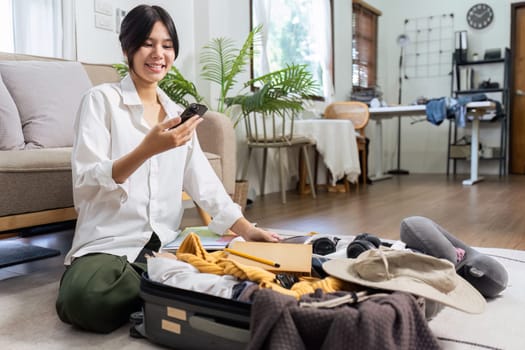 Young pretty traveler woman using smartphone to book a hotel while preparing travel suitcase.