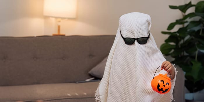 Children cosplay as white sheet ghost with sunglasses holding a bucket of pumpkins for Halloween.