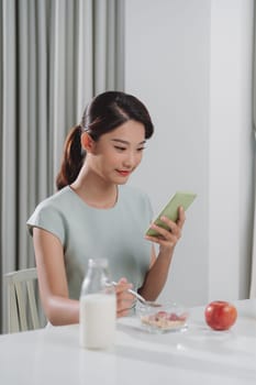 Smiling pretty asian young woman surfing on Internet while having healthy breakfast at home