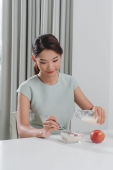 Smiling young woman pouring milk into a bowl while sitting and having breakfast 