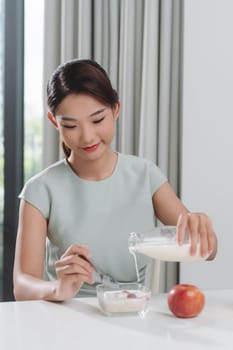 Happy  woman pouring fresh milk from glass into bowl with cereals, having delicious meal