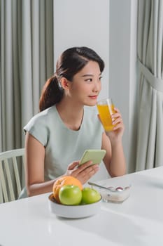 Young joyful woman drinking orange juice while holding mobile phone sitting near a kitchen table