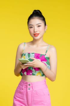 Smiling asian woman holding smartphone and looking at the camera over yellow background
