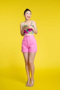 Smiling asian woman holding smartphone and looking at the camera over yellow background