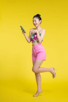 Portrait of a smiling young asian woman using mobile phone isolated over yellow background