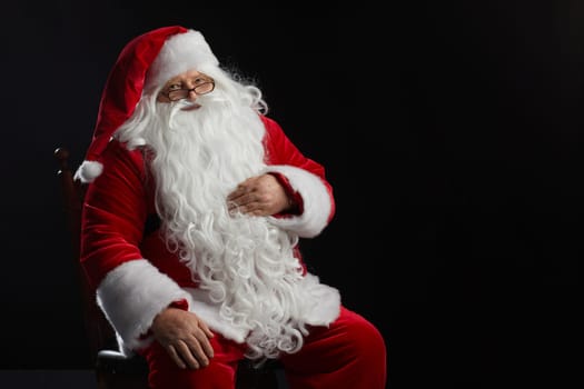 Santa Claus sits on a black background and looks at the camera. Christmas, New Year, Holiday Celebration, space for text