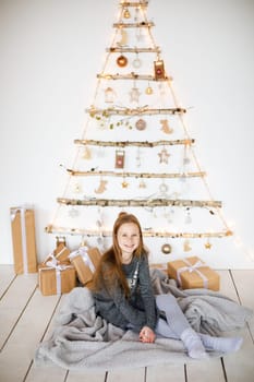 little girl holding craft gift against background of christmas tree with wooden new year toys hanging on wall, vertical photo