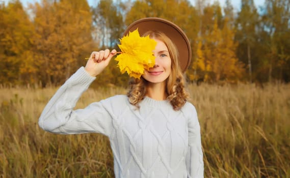 Autumn portrait of happy young woman with maple yellow leaves, cheerful girl smiling wearing hat, sweater outdoors in park, warm fall season