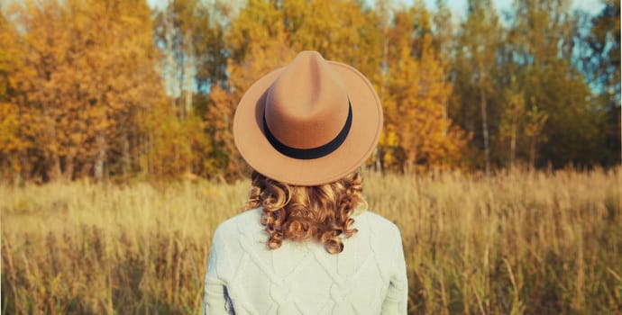 Back view of beautiful happy young woman in brown hat enjoying warm weather, looking at landscape in autumn park with yellow leaves outdoors, fall season, nature background