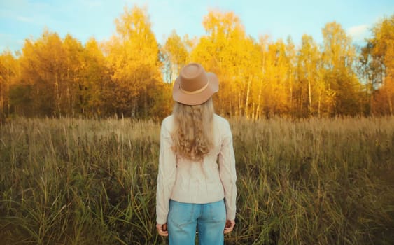 Back view of beautiful happy young woman in brown hat enjoying warm weather, looking at landscape in autumn park with yellow leaves outdoors, fall season, nature background