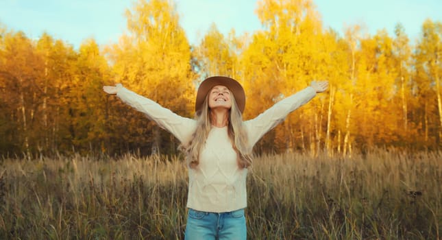 Happy smiling woman in autumn park raising her hands up, joyful girl in hat, sweater enjoying warm sunny weather, fall season, nature background