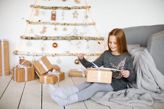 little girl holding craft gift against background of christmas tree with wooden new year toys hanging on wall