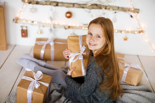 little girl holding craft gift against background of christmas tree with wooden new year toys hanging on wall
