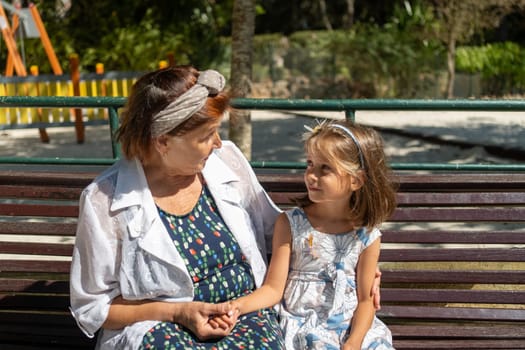 Senior woman and cute girl sit on park bench, gazing at each other with warm smiles, sharing tender and affectionate moment during sunny day