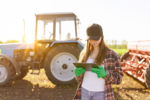 Farmer woman with tablet working in field. Concept of technology in agriculture. Harvest season in an organic farm on a bright sunny day. Tractor at work.