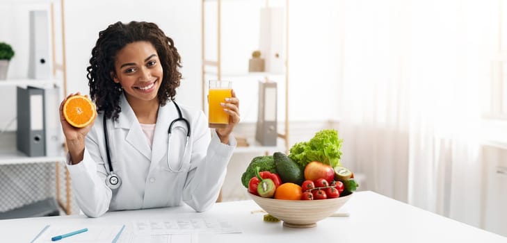 Cheerful female nutritionist african woman holding fresh orange juice and orange half in her hands while posing at modern clinic, healthy lifestyle concept