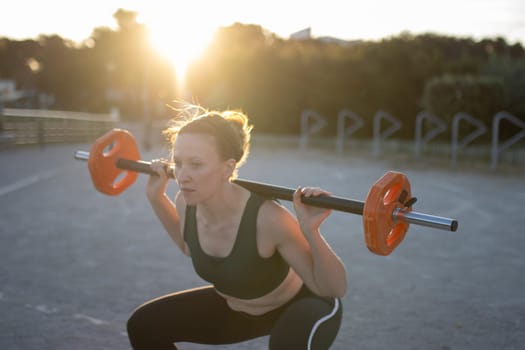 Young woman is doing squats while lifting weights outdoors