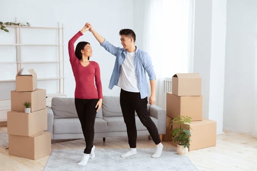 Celebrating Moving Day. Happy young asian tenants dancing among unpacked boxes in new home