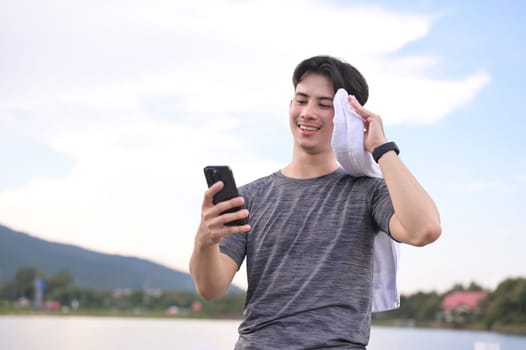Smiling young male runner resting and using mobile phone in the park after morning run.