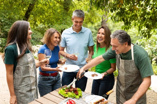 A group of five friends enjoys a lively barbecue in a garden, sharing food and laughter. They hold drinks and plates, celebrating their time together on a sunny day.