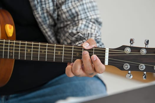 Close up of a man in plaid shirt playing an acoustic guitar on sofa at home. Leisure, hobby and activity concept.