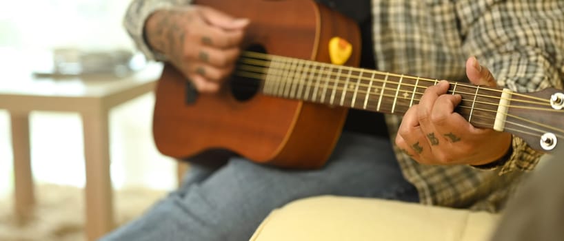 Young man playing an acoustic guitar while sitting on sofa at home. Leisure activity and hobby concept.