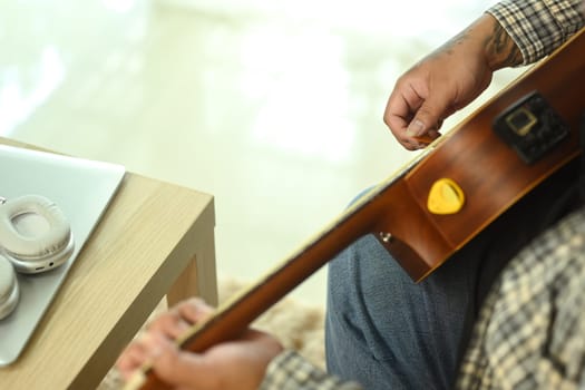 Young man playing an acoustic guitar while sitting on sofa at home. Leisure activity and hobby concept.