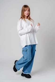 A joyful young girl with long hair wearing a white sweater stands against a plain background close up