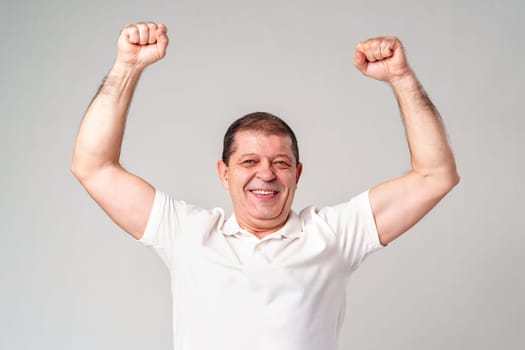 Happy older man showing off his strong biceps while standing against a light gray background close up