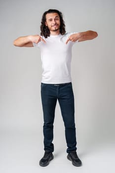 A young man with curly hair joyfully pointing down while wearing a plain white t-shirt against a neutral background close up