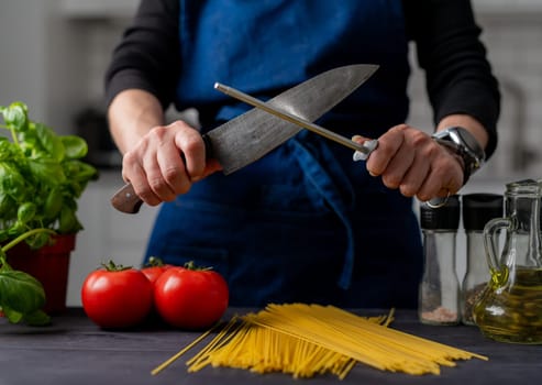 Large Kitchen Knife And Sharpener In Female Hands Are In Close-Up Over Table With Spaghetti, Tomatoes, Spices, And Basil