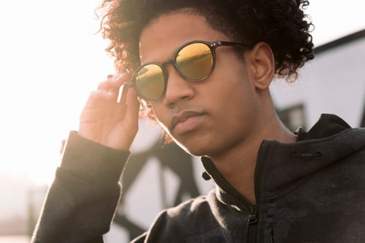 confident curly haired african american male teenager adjusting trendy sunglasses while standing against blurred urban background