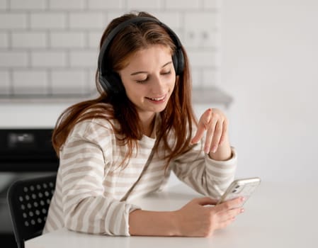Cheerful Girl Listens To Music On Headphones Through Her Phone At Home In The Kitchen