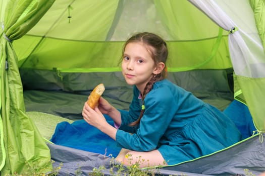 A young girl relaxes in a green camping tent, enjoying a delicious snack and natures beauty with a serene smile