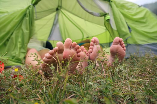 Two pairs of bare feet are comfortably relaxing inside a tent pitched on a grassy meadow while enjoying nature